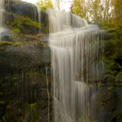 Cascada en los cañones del río Lozoya, con rocas cubiertas de musgo y hojas secas. El agua cae en velos blancos sobre la piedra oscura.