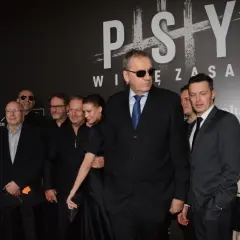 Grupa aktorów z filmu "Psy" pozuje do zdjęcia. W centrum mężczyzna w okularach przeciwsłonecznych i garniturze.