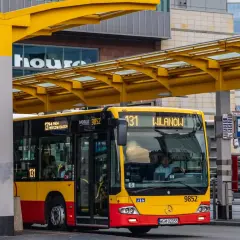 Kierowca autobusu linii 131 dojeżdża do przystanku Wilanów. Żółto-czerwony pojazd czeka na pasażerów pod żółtą wiatą.