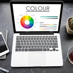 Na ekranie laptopa widać koło barw i palety kolorów, prezentujące różnice między cmyk vs rgb. Obok telefon, notes i filiżanka herbaty.