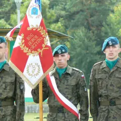 Trzech żołnierzy rezerwy w mundurach i beretach stoi obok sztandaru.