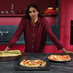 Mann in bordeauxrotem Hemd präsentiert frisch gebackene Pizzen. Im Hintergrund ein Ofen mit Pizzafunktion, der eine weitere Pizza bäckt.