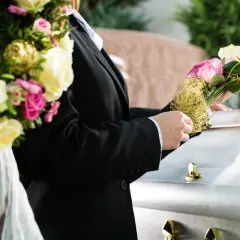 Osoba w czarnym garniturze kładzie bukiet róż na białej trumnie.