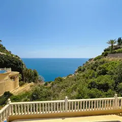 Vista desde una terraza con barandilla blanca hacia el mar azul, con villas en laderas verdes. La puerta del mar San José se abre a un paisaje idílico.