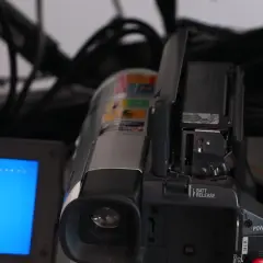 En videokamera med öppen lucka och en blå skärm. Kablar syns i bakgrunden, en påminnelse om analoga bilder och äldre teknik.