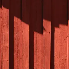 Röd träfasad med skuggor som faller som vertikala streck. Träet är målat tunt, vilket ger en rustik känsla.