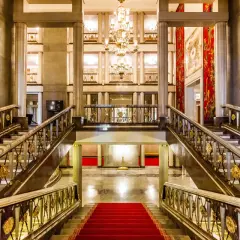 Elegancki foyer z marmurowymi schodami, złotymi balustradami i kryształowym żyrandolem. Czerwony dywan prowadzi do przestronnego holu.