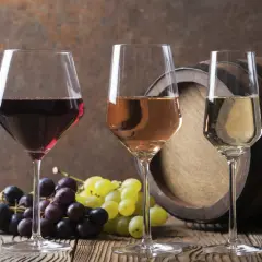 Trzy kieliszki z winem: czerwonym, różowym i białym, obok kiści winogron i beczki. Idealne na wieczór z **wino bezalkoholowe**.