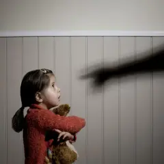 Mała dziewczynka z pluszakiem patrzy z przerażeniem na cień ręki wskazującej palcem. To symbol przemocy domowej.