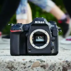 Nikon D500 na kamieniu, z rozmazanymi nogami w tle. Chcesz wiedzieć, jak sprawdzić przebieg aparatu Nikon?