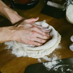 Dłonie delikatnie rozciągają ciasto na pizze, tworząc idealny okrąg na drewnianym blacie. Mąka rozsypana wokół sugeruje, jak rozciągnąć ciasto na pizze.