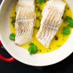 Dwa filety halibut pieczony ze skórą w maśle z ziołami i kolendrą, podane w białej misie z czerwonymi uchwytami.