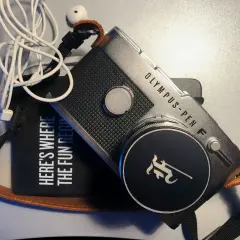 The Olympus Pen FV camera, with its iconic design, sits beside a black pouch that reads "HERE'S WHERE THE FUN BEGINS." White earbuds are tangled nearby.