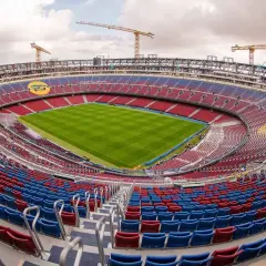 Camp Nou w Barcelonie w trakcie modernizacji. Widok na pusty stadion z zieloną murawą i czerwono-niebieskimi trybunami, nad którymi górują dźwigi budowlane.