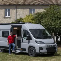 Un homme pose devant un **fourgon ou camping car** gris, porte ouverte, devant une maison en pierre et des arbres.