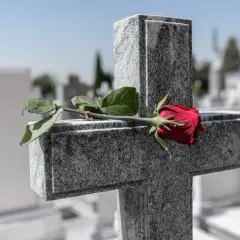 Czerwona róża na kamiennym krzyżu. Cichy symbol pochówku bez pogrzebu, samotne pożegnanie na cmentarzu.