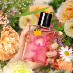 Dłoń trzyma flakonik z różowym płynem i symbolem Wenus zwieńczonym koroną. Czy perfumy z feromonami działają? Otoczenie bujnych kwiatów sugeruje zapach.