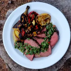 Skivad picanha med perfekt picanha temp, toppad med chimichurri och grillade grönsaker. Serveras på en vit tallrik på en trädstam.
