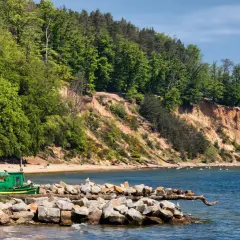 Zielona łódź rybacka przy kamienistym brzegu, na tle zielonego lasu i piaszczystej plaży. W oddali widać wysokie, strome urwisko porośnięte drzewami.