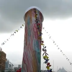 Un'installazione colorata, alta e decorata con festoni di fiori. Si immagina di poter toccare il naso di Pulcinella.