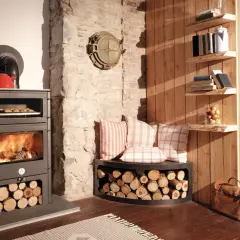 Gemütlicher Raum mit brennendem Kaminofen, darauf eine rote Teekanne. Daneben ein Holzstapel und Kissen. Ein weidenholz Kamin sorgt für Wärme.