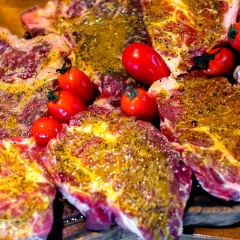 Soczyste plastry karkówki, marynowane i przyprawione, gotowe do grillowania. Obok leżą czerwone pomidorki koktajlowe.