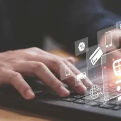 Dłoń na klawiaturze laptopa, nad nią ikony symbolizujące zakupy online: koszyk, karty płatnicze, dostawa. Sukces każdego ecommerce retailer.