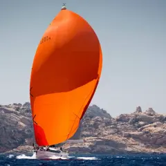 Una barca a vela con una grande vela arancione sfreccia sull'acqua blu, con rocce frastagliate sullo sfondo.