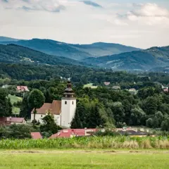 Widok na wieś z kościołem i domami, otoczoną przez zielone wzgórza i lasy. W oddali widać łagodne **górki wielkie**.