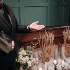 Mężczyzna w garniturze prezentuje trumnę, trzymając książkę. Scena sugeruje pogrzeby z zasadami, z kwiatami i świecami tworzącymi uroczystą atmosferę.