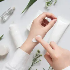 Eine Hand testet eine weiße Creme, umgeben von Pflegeprodukten und zarten Blüten. CI 45410.