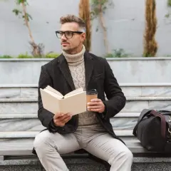 Elegancki mężczyzna w okularach, z książką i kawą, siedzi na ławce. Jego styl emanuje klasą i old money.