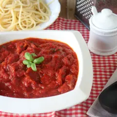 Un tazón blanco lleno de salsa marinara casera, decorada con albahaca fresca, junto a espaguetis cocidos.