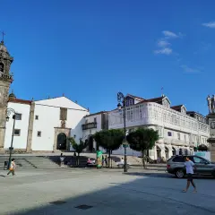 La plaza Betanzos, con su iglesia de torre campanario, edificios blancos y un monumento, bajo un cielo azul.