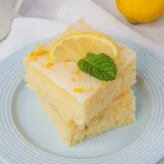 Zwei Stücke zitronenkuchen mit joghurt, garniert mit Zitronenscheibe und Minze, auf einem hellblauen Teller.