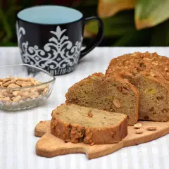 Un delicioso bizcocho de plátano, cubierto de frutos secos, acompañado de cacahuetes y una taza.