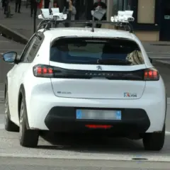 Voiture blanche avec équipement de **voiture radar embarqué** sur le toit, circulant dans une rue de Lyon.