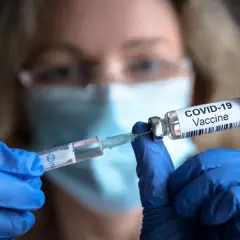Osoba w niebieskich rękawiczkach przygotowuje szczepionki na covid, pobierając płyn do strzykawki z fiolki z napisem "COVID-19 Vaccine".