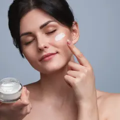 Frau trägt eine Tages- und Nachtcreme auf. Sie hält ein Glas mit der Creme in der Hand und tupft sie mit dem Finger auf ihre Wange.