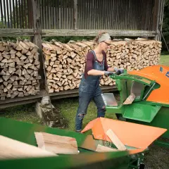 Frau bedient eine Schiebetisch Kreissäge für Brennholz, um Holzscheite zu schneiden. Im Hintergrund sind Holzstapel zu sehen.