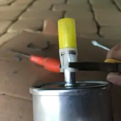 Dłoń trzymająca szczypce przy filtrze paliwa z żółtą zatyczką.