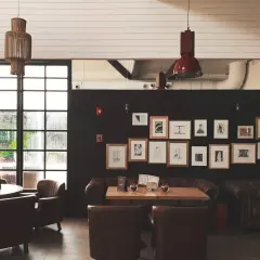 Una lámpara de yute cuelga sobre mesas y sillones de cuero en un café con paredes oscuras y una gran ventana.