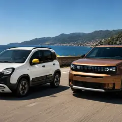 Due Fiat Panda 2026, una bianca e una color rame, sfrecciano su una strada costiera panoramica con il mare e le montagne sullo sfondo.
