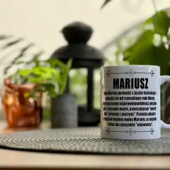Biały kubek z napisem "MARIUSZ" i opisem pochodzenia imienia. W tle rośliny i ozdobna lampa.