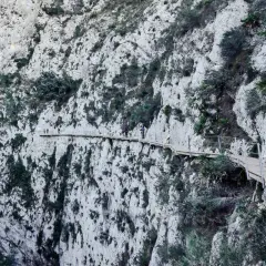 Personas caminan por la **ruta de las pasarelas**, un sendero de madera colgado en un acantilado rocoso con vegetación.