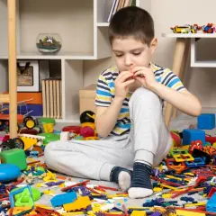 Chłopiec siedzi pośród rozrzuconych zabawek, próbując posprzątać swój pokój. Wszędzie widać klocki, samochody i pluszaki.