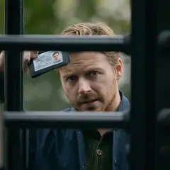 Ein Mann mit blondem Haar und Bart hält einen Ausweis hoch, während er durch ein Gitter blickt. Spannung in Slow Horses Staffel 3.
