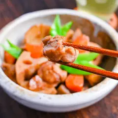 A close-up of a bowl of chikuzenni recipe, with chopsticks lifting a piece of chicken. The dish contains chicken, lotus root, carrots, and green beans.