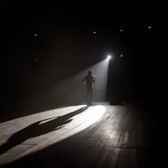Mężczyzna stoi na scenie w świetle reflektora, gotowy do występu na open mike stand up. Długi cień rzuca się na drewnianą podłogę.