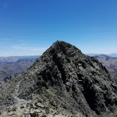 Cima rocosa de montaña con excursionistas y vista panorámica de picos bajo un cielo azul.